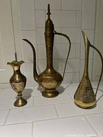 Three vintage brass vessels on white tiled surface showing two ewers and one decorative vase with colored detailing.