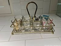 Photo showing a set of six vintage shot glasses placed in a brass-tone metal rack holder with a handle on a tiled surface. The glasses have varied designs including prints and logos in black, red, and blue.