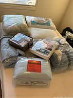 Overall view of bedding items on bed including comforters, blankets, sheets, pillows, and mattress cover packaged mostly in plastic wrapping.