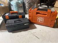 Three toolboxes/cases on concrete floor: one orange Fein Multimaster case, one black and orange taken toolbox, one black Holt toolbox.