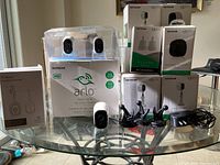 Overview of the Netgear Arlo security cameras and accessories on glass table, including 2-pack camera box, single camera, mounts, power adapter, and other accessory boxes.