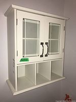 Front angled view of the white wall mounted storage cabinet with two glass doors and three cubby compartments below.