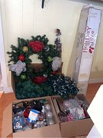 Artificial wreath decorated with red mesh bows and ornaments beside two cardboard boxes with assorted Christmas ornaments and string lights, and a tall box labeled 'Holiday Time' containing an artificial Christmas tree.