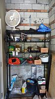 Metal shelving unit holding varied items including a white Larko fan on the top shelf, fertilizer bucket on bottom shelf, and various gardening and hardware supplies.