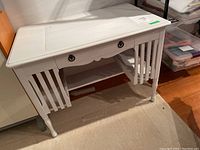 Front angled view of the white wood desk showing drawer, open bottom shelf, plastic writing pad on top.