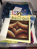Clear plastic bin with assorted quilting fabrics, quilting magazines including Bargello Quilts and quilting guides, organized with fabrics underneath.
