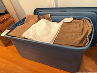 Large blue plastic storage bin open showing various folded fabrics inside including brown, white, and plaid pattern.