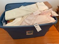 Blue plastic storage bin filled with assorted fabric pieces, including a smooth white satin-like fabric and pink striped fabric, loosely folded inside the bin.