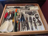 Overview of the open drawer containing assorted flatware, kitchen knives with colored handles, pizza cutters, and a white porcelain double-section spoon rest.