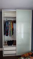 Front view with left door open showing clothing rod, hanging garments, shoes on shelf, and two drawers below.
