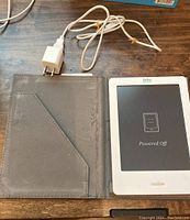 White Kobo e-reader inside a grey protective case with white charger cable and plug. Device screen displays powered off status.