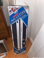Box of Logan Compact Mat Cutter model 301-M standing upright against a wall, showing branding and product images.