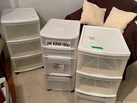 Photo showing three white plastic drawer organizers side by side on floor, each with three drawers and casters. One organizer has a sticker on top drawer front.