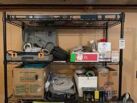 Full view of three metal shelves filled with various tools and supplies including boxes, a clock, cleaning rags, and hardware.