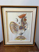 Front view of framed artwork showing stylized rooster and human faces.