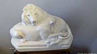 Front view of the white glazed ceramic sculpture showing the recumbent lion couple with clear details on the lions' facial features and the base with artist signature.