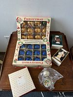Two sets of boxed vintage glass Christmas balls in gold and blue colors with 12 ornaments each, plus some loose silver glass balls with painted decorations, and two holiday-themed books on a wooden table.