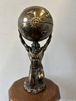 Front view of Atlas kneeling with muscular torso and beard, holding large zodiac globe overhead on a wooden surface against plain background.
