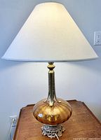 Single amber glass table lamp with off-white fabric shade, gilt metal base with ornate details, placed on wooden surface.