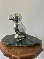 Polished metal puffin bird sculpture with smooth finish on natural stone base, photographed front-facing on wooden surface.
