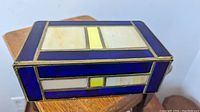 Angled view of the closed stained glass jewelry box on a wooden surface showing blue, yellow, and cream stained glass panels with brass framing
