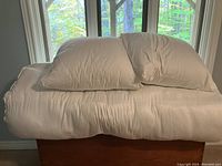 Photo showing two standard pillows and a rolled double mattress cover/topper stacked on a wooden chest in front of windows with forest view.