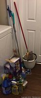 Photo showing assortment of cleaning bottles, mop buckets, mop handles, dustpans, and brooms against wall.