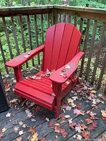 Red Adirondack chair in outdoor setting on wooden deck, sitting upright with fallen autumn leaves around and on it.