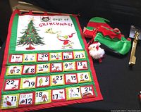 Full view of the Grinch advent calendar wall hanging with 25 numbered pockets, elf hat, booties, gold wreath hook, and Santa door knob cover displayed on black surface.
