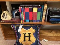 Multiple Harry Potter books and DVDs arranged on a wooden shelf, including an unofficial cookbook and a film wizardry book, also a themed cushion placed below the shelf.