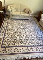 Full view of the large floral area rug beneath a couch, showing lattice diamond pattern with pink and blue flowers on a cream background and tassels on the edge.