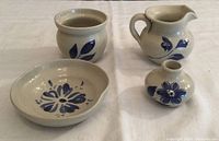 Four small Williamsburg pottery pieces together on a white cloth. Includes a bowl, pitcher, small vase, and jar, all beige with blue decoration.