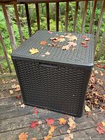 Black outdoor storage box with rattan wicker pattern, shown closed with leaves on top on wooden deck outside.