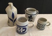 Four Williamsburg Pottery stoneware pieces with blue hand-painted leaf designs against a beige background, including mugs of varying sizes, a small handled vase/utensil holder, and a jug with an open front for candle placement.
