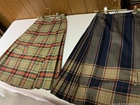 Two wool skirts laid out side by side showing pleated design, different plaid patterns and colors, displayed flat on a white surface.