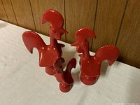 Three red metal rooster figurines together on a white cloth surface against a wood panel background, showing different heights.