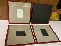 Four wooden picture frames placed on a wooden floor against a wood-paneled wall. Two frames show their glass sides with mats inside, two show the backs with hanging hardware visible for mounting, all frames appear square with approximately 17x17 inch outer dimensions.