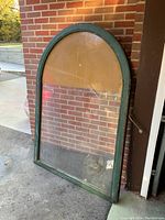 Two photos showing each of the tall arched mosaic stained glass windows leaning against a brick wall, with green painted wooden frames and some wear on the reflectiveness and paint.