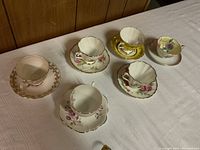 Six vintage teacups and saucers arranged on a table showing floral and gold decorative patterns with varying colors and styles.
