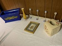 Blue planter, yellow vase, floral picture frame, floral ceramic planter, and decorative hook rack on white cloth-covered surface against wood paneling.