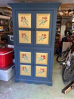 Front view of a tall solid wood cabinet painted dark blue with yellow panels on the doors featuring red and blue flower hand-painted designs.
