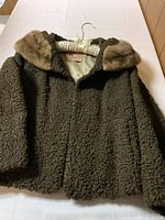 Front view of dark brown textured fur coat with light brown fur collar hanging on a white padded hanger on a bed