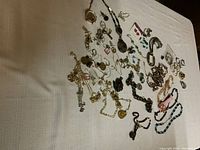 Multiple pieces of costume jewelry scattered on a light cloth surface, showing variety of necklaces, bracelets, earrings, and two wristwatches.