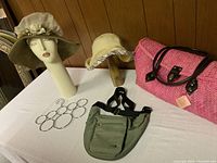 Wide shot of the two hats, pink woven handbag, green waist bag, and metal accessory holder on a white table with wooden background