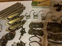 Top-down view of various vintage hardware including brass and metal drawer pulls, round knobs, wooden knobs, and polished metal hooks arranged on a white surface.