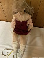 Front seated view of life size porcelain doll with burgundy dress and white socks and shoes, placed next to a measuring tape showing approx 35 inches tall