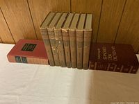 Photo shows 7 volumes of The Book of Knowledge with tan hardcover and brown spines, along with a maroon Standard Desk Dictionary and the Familiar Quotations book, all arranged upright on a white cloth surface against a wood-paneled background.