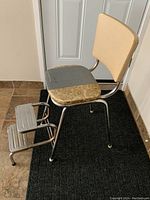Vintage metal step stool chair with worn upholstery and attached two steps, seen from the side in front of a white door on tile floor with black mat.