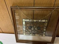 Framed stained glass picture featuring floral pattern with a hanging metal chain attached.