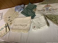 Wide view of assorted vintage linens including pillowcases, napkins, and table runners in various embroidery and lace designs on cream, white, green and blue fabrics.